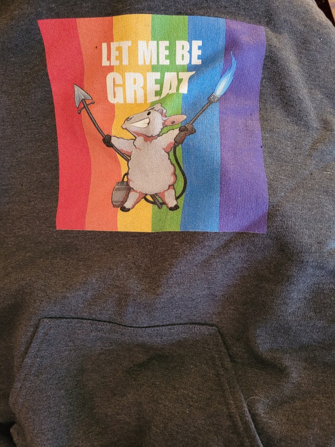 Hoodie with an illustrated sheep carrying a spear and a flamethrower on a rainbow background saying "Let Me Be Great"