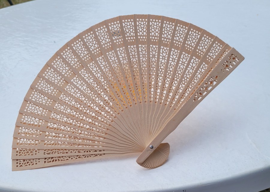 A wooden hand fan with cutouts along the blades, leaning up against a cup of wine you can just see through the cutouts