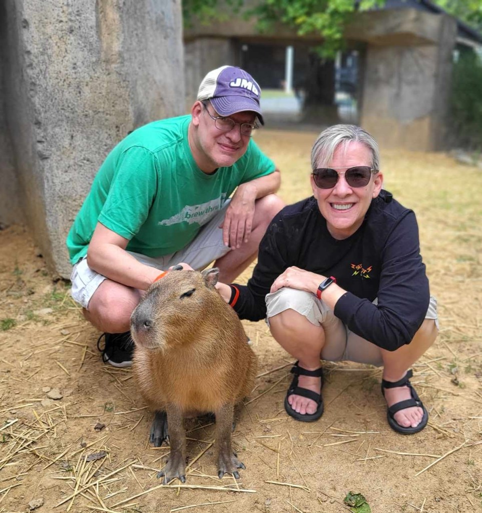 A photo of my brother & myself kneeling next to a capybara named Tater. Tater is 7 months old and weighs approximately 35 pounds. He is shaped somewhat like a potato with legs. His hair is medium brown and very coarse. He has dark eyes that with markings around them that look like cateye eyeliner. His ears are small and round and can flatten down when swimming. His feet have 3 toes that are partially webbed, also for swimming.