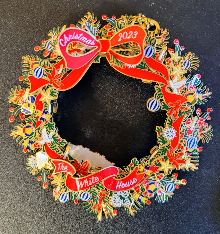 The 2023 White House Christmas ornament. It is a metal wreath with a red ribbon and lots of little painted baubles on it.