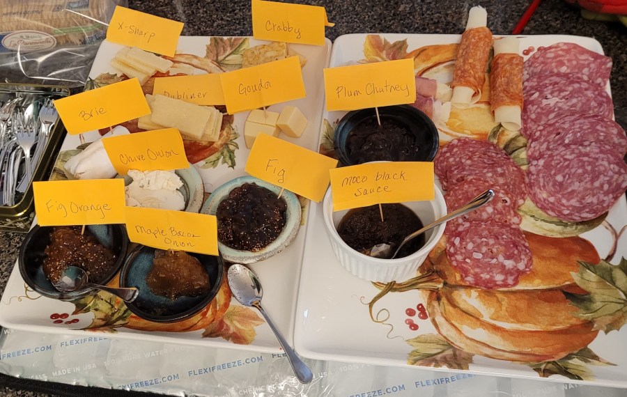 A photo of two thanksgiving themed platters with an assortment of cheeses, salamis, and accompaniments on them. With post-it notes on toothpicks identifying what is what