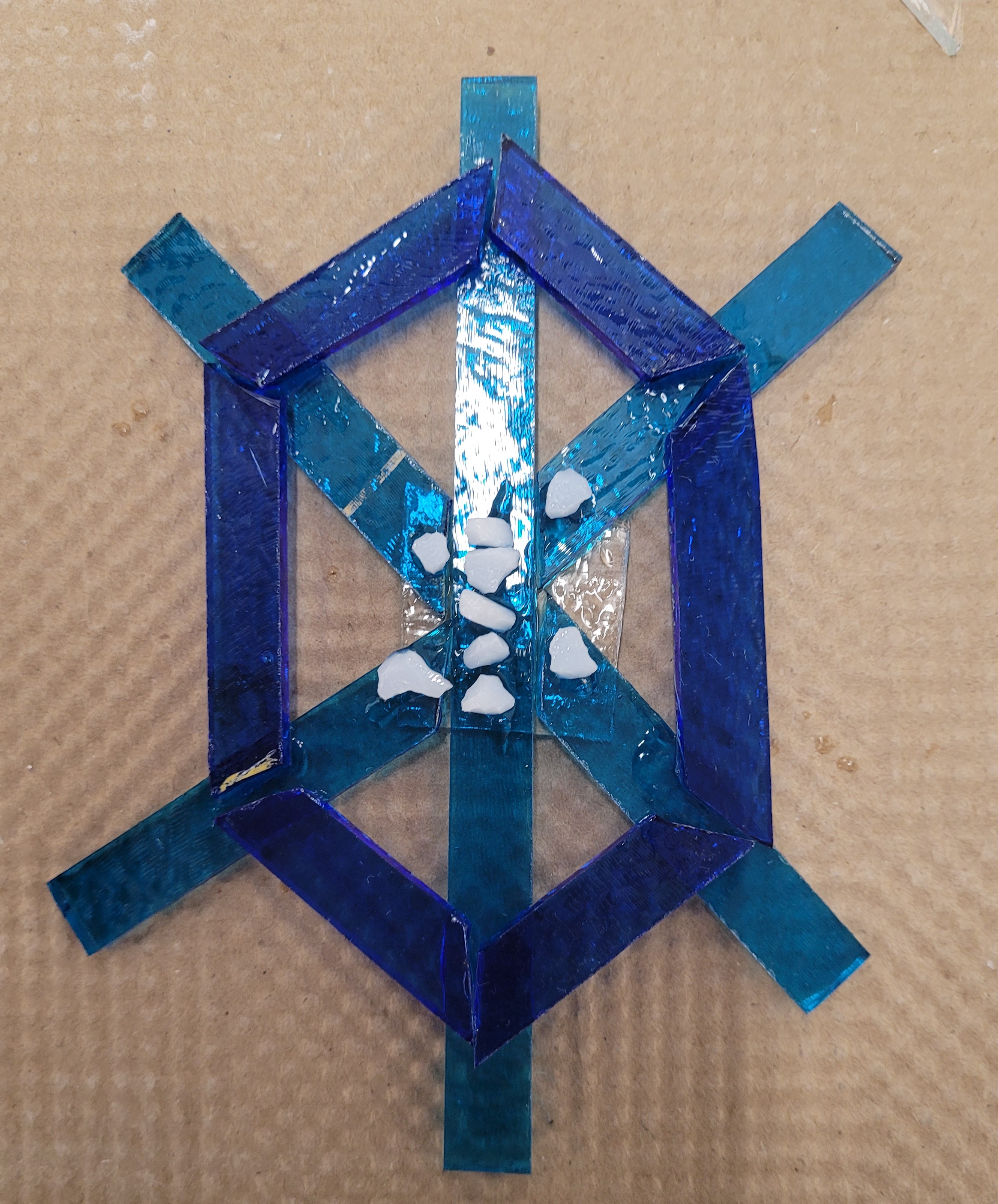 An un-fired 6 pointed snowflake. The points are strips of sky blue glass. Then there are royal blue strips connecting the points at about the halfway points of the main glass strips. There is also a bit of white frit (tiny bits of glass) in the center. It is more turtle than snowflake shaped.