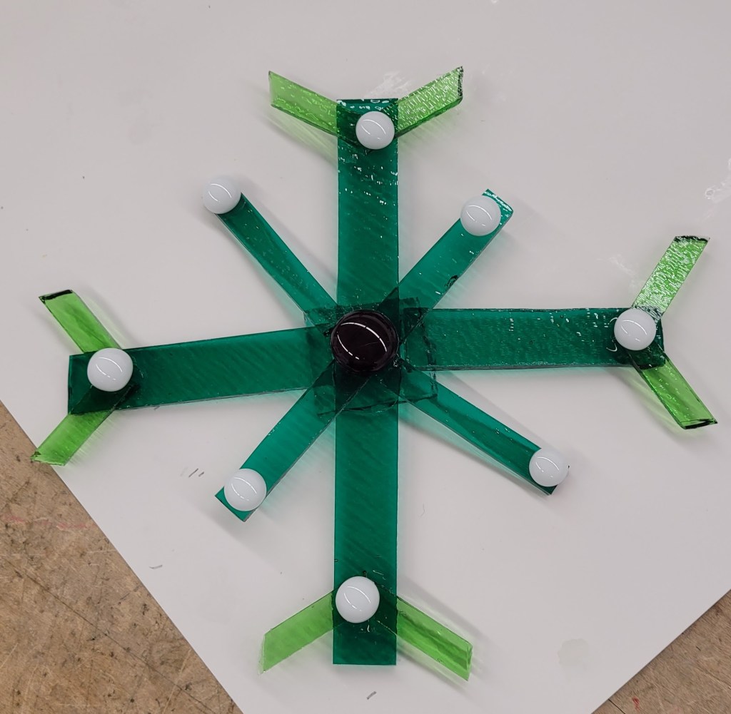 An un-fired glass snowflake. It is green with 8 points. 4 long points horizontal and vertical, and narrower points diagonally. At the end of the long points are two lighter green narrow pieces of glass forming an arrow towards the center. At the end of all 8 points is a white glass button, and there is a dark red button in the center.