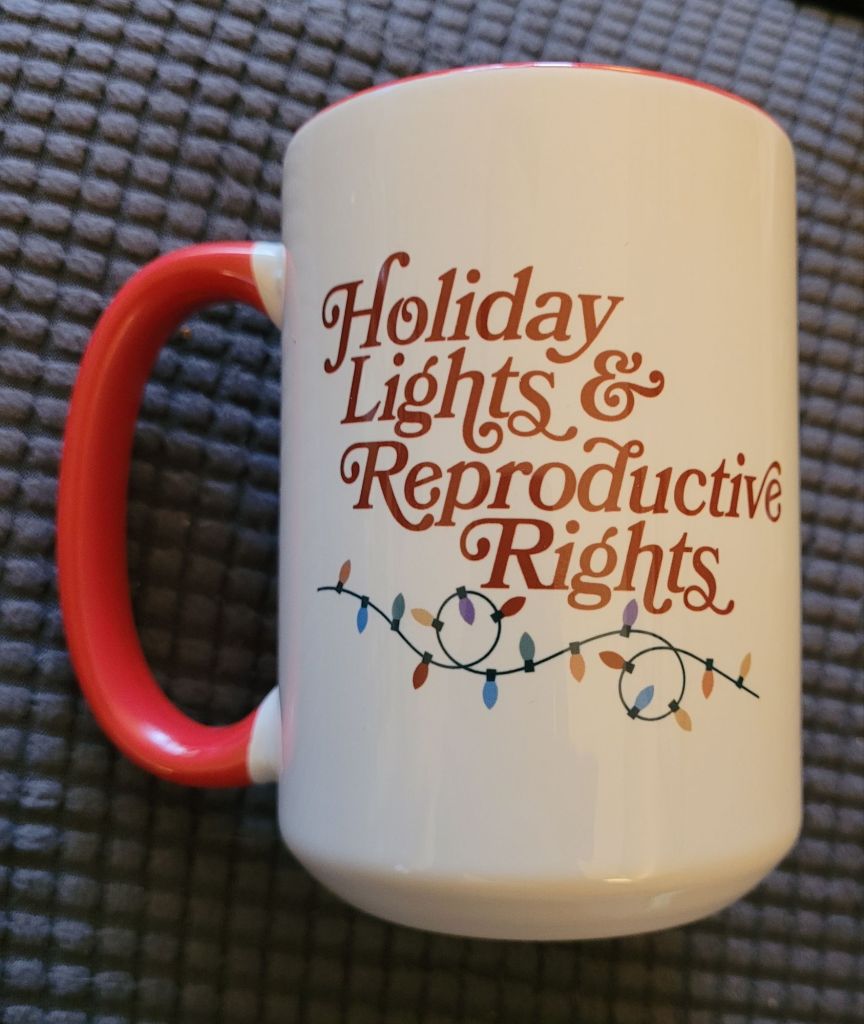 A white coffee mug with a red handle, with "Holiday Lights & Reproductive Rights" written on it with a string of lights underneath.