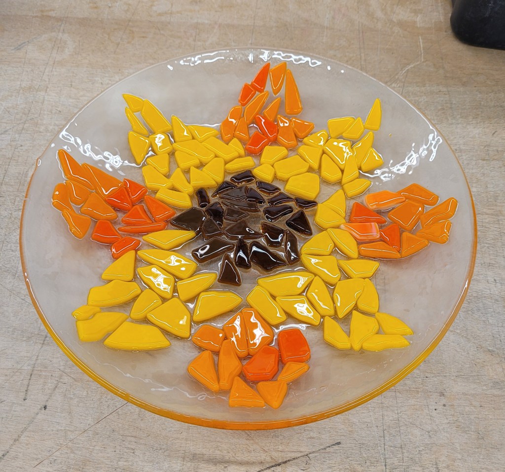 A fired circle of clear glass, with a mosaic sunflower on it. The center of the sunflower is opaque and transparent brown bits, and the sunflower petals are opaque yellow, and opaque orange. It got a tack fuse so the texture of the mosaic pieces is still there. It was flat and has now gotten a slump firing in a mold, so it is a bowl now.