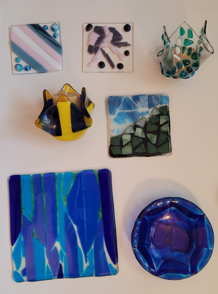 A variety of fused glass pieces, from top to bottom, left to right: A fired suncatcher. It has 5 diagonal color strips - blue, pink, white, pink, and blue - the colors of the trans pride flag. In the other corner there are three blue cabochons (dots). A fired suncatcher. A clear plate with a pink and purple jellyfish (or vaguely jellyfish shaped design) - it is very mosaic-y, the body has lots of pieces of pink and purple glass. There are ocean-ish blue cabochons around it. A fused glass votive candle holder. It started with a flat square of fused glass, placed over a round mold to make the bottom and in the second firing, it slumps down into wavy glass along the sides. The glass itself is clear with with small bits of blue, blue-green, green, and white glass on top that spread out from the middle to the corners, giving an ocean wave feel to it. A fused glass votive candle holder. It started with a flat square of fused glass, placed over a round mold to make the bottom and in the second firing, it slumps down into wavy glass along the sides. It is a clear square of glass that is covered with large pieces of royal blue, yellow, medium blue, and very light yellow glass. A strip of royal blue runs from one corner to the other. There are bright yellow strips on either side of it. Then there are strips running perpendicular in the bright yellow and royal blue, with some medium blue on upper left, and then the very pale yellow is at all four corners. A fired piece of 5x5 inch glass. In the lower half, there are pieces of green glass going upwards towards the upper right hand side, and coming down from the left are pieces of streaky blue and white glass. The blue and white glass is surrounded with white frit, and the green is surrounded with black frit. The frit is like sand and got kinda messy and all over the place. A fired light leak piece. It is varying shades of blue shards up and down with space in between. In the lower half, the spaces are filled with green frit (ground glass) and in the upper half, the spaces are filled with pale yellow frit. The yellow frit came out almost clear - which is OK! I was feeling “ocean” with all the blues and wanted the green at the bottom as “sea kelp” and the yellow at the top as light filtering through, and it works! A circular plate of fused glass. It was made from chunks of a pattern bar of layers of blue and clear glass - it is several shades of blue and I love it.