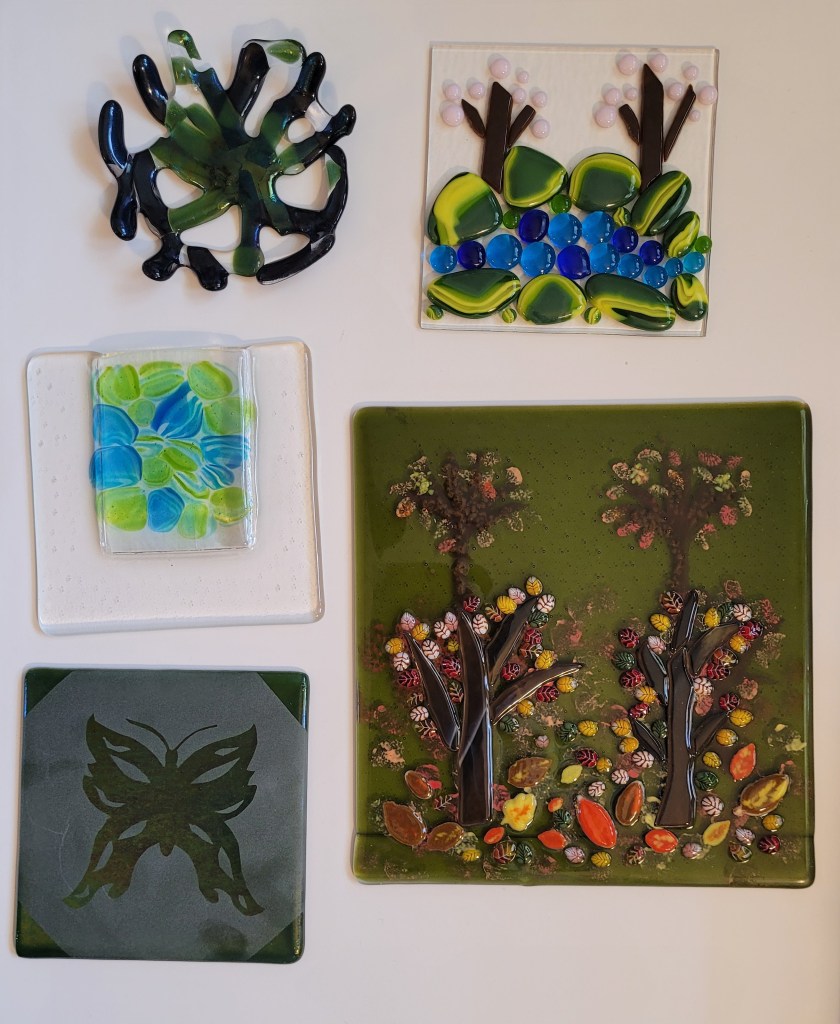 Several fused glass pieces. Top to bottom, left to rightL A "coral bowl" - made with strips of glass and is reminiscent of ocean coral. The various strip colors are olive green rainbow iridescent (it changes from greens to blue-purples depending on the light) then blue adventurine, which is a sparkly navy blue, and then clear bits for stability here and there. A fired piece of 5x5 glass with a landscape. The landscape is green & yellow cabochons (big dots) for grass at the bottom. Then there are blue transparent dots of various shades for water. Then more of the green & yellow cabochons for more grass above the water. Then two trees of brown glass with pink dots around them as cherry blossom trees. It has been ”tack fused" so the glass is still raised up on the base plate, but soft and rounded. A "pocket vase." It is a sheet of 6x6 inch clear glass, with fiber paper in the top center, about 3 x 4 inches. There is another sheet of glass on top, which will melt down around the fiber paper, leaving a hole at the top. On the very top, I have added transparent blue and green cabochons. The fiber paper in the top center has been removed after firing, and there is a pocket at the top where flowers can go. On the very top, I have added transparent blue and green cabochons, the cabochons have clear stripes in them and look like flowers themselves now that they have "melted" into the glass. A sandblasted piece. Contact paper was put on the corners and in the center in a butterfly shape. Then the exposed glass was sandblasted, giving it a matte appearance. The contact paper is removed, leaving the pattern and shiny glass. It is green iridescent glass. A multi layer landscape. It is on transparent fern green glass. The trees in the background are painted with some additional frit for texture. The trees in the foreground are glass, with the leaves made of murrine and freeze and fuse.