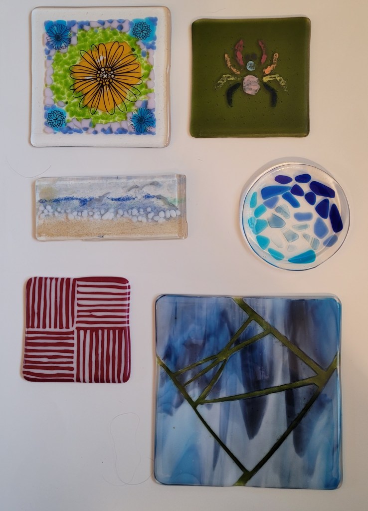 A selection of fired glass pieces. From top to bottom, left to right: A 6x6 fired glass plate. In the center are bits of yellow glass in a sort-of circle. That is surrounded by green coarse frit (ground glass bits) then some small bits of pink surrounding that, then blue pieces in the corners in smaller circles, and then more blue coarse grit around the edges. The high temp firing has smoothed everything out. There are decals on it of line art in various flower shapes. There is a large flower over the center section of yellow, and small flowers over the blue parts of the corners. A piece of 5x5 inch green transparent glass, with a stenciled spider painted onto it over it. The legs and body have been painted in with various colors of glass paint, including red, orange, yellow, blue, pink, turquoise, and purple. A three layered rectangular piece - the bottom layer is ocean & sky, the middle layer, the sun & seagulls, and sand on the top layer. A fired circle of clear glass about 5 inches across, with bits of transparent blue glass of varying shades in a mosaic pattern. It had a full fuse firing, and is flat and the blue bits are very smooth embedded in the clear glass - it then had a slump firing in a mold and is now a shallow dish A square "strip construction" piece. Strips of red and cream glass are turned on their edge and sandwiched together with the red and cream alternating. There are four blocks of the strips, the upper left and lower right have the strips running vertically, and the other two blocks have the strips running horizontally. This shot is from the top, and it is very smooth and shiny. An 8x8 inch piece of fired glass. The main color is a streaky blue and white opaque glass that was about 7 1/2 by 7/12 inches, broken into large chunks and placed on an 8x8 inch clear sheet, so that there is space between the broken pieces. In those gaps, it has been filled in with fine frit, which is basically very tiny pieces of glass, almost like coarse sand. The frit is light green and is transparent and the light "leaks" through it. It's in a windowsill with the sunlight coming through so the “light leak” is more apparent.