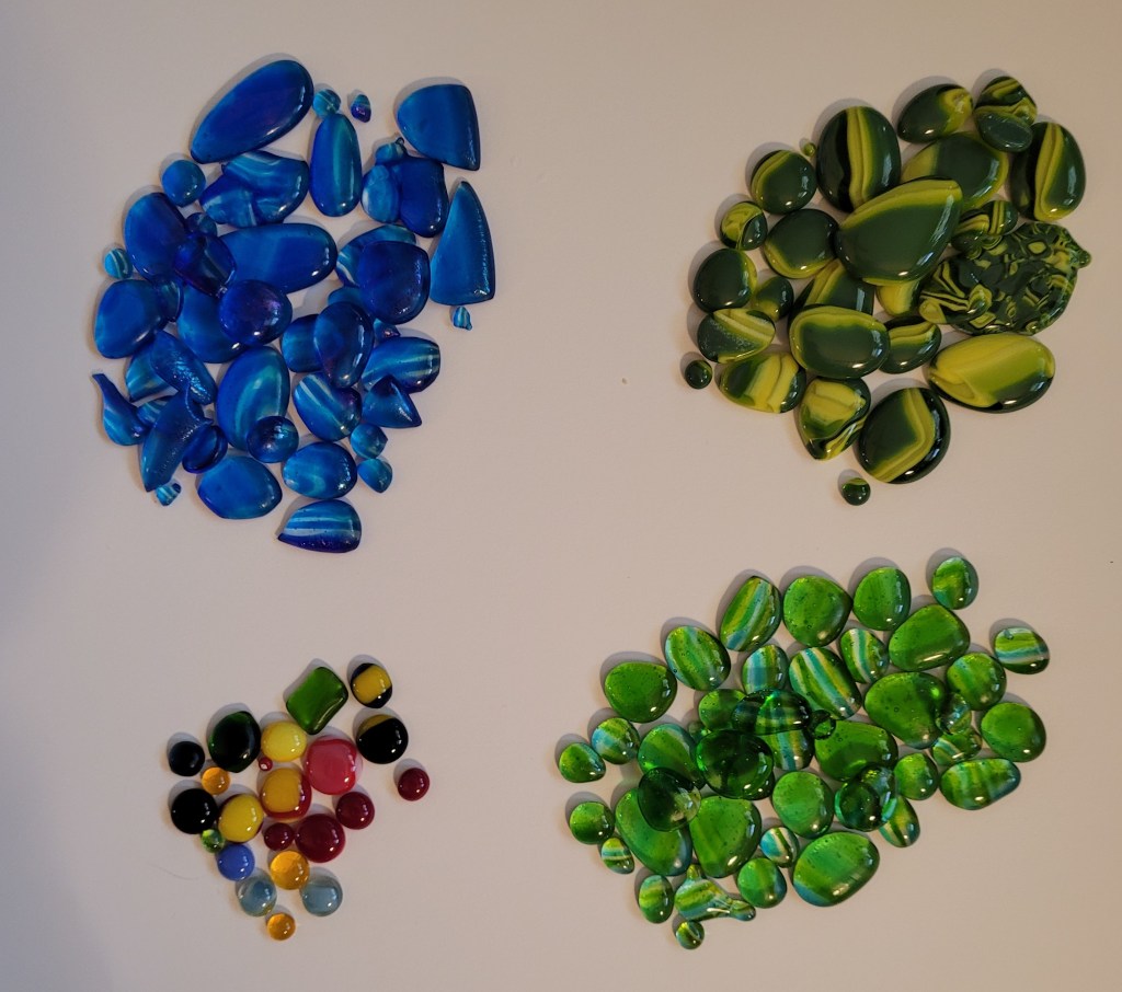 4 piles of cabochons/glass dots. Upper left are made from layers of transparent blues and clear glass. Upper right, dark green and yellow glass. Lower left a variety of small glass dots in different colors. Lower right made from layers of transparent greens and clear glass.