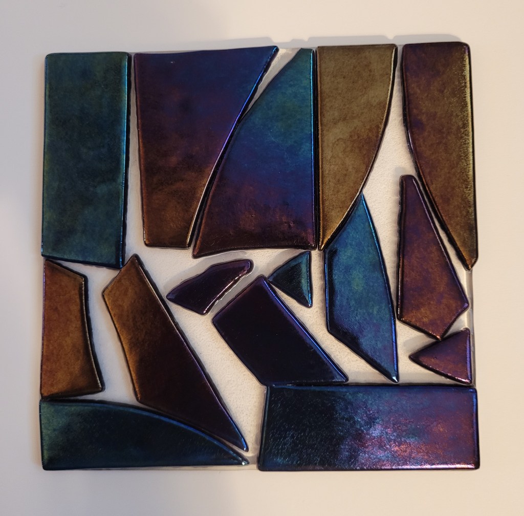 An fired piece of 5x5 inch glass. It is a clear glass base with large chunks of dark iridescent glass arranged on top of it in a mosaic-ish fashion. The iridescent pieces shine in blue, purple, and gold - depending on the piece of glass and how the light is hitting it. It has been ”tack fused" so the glass is still raised up on the base plate, but soft and rounded.