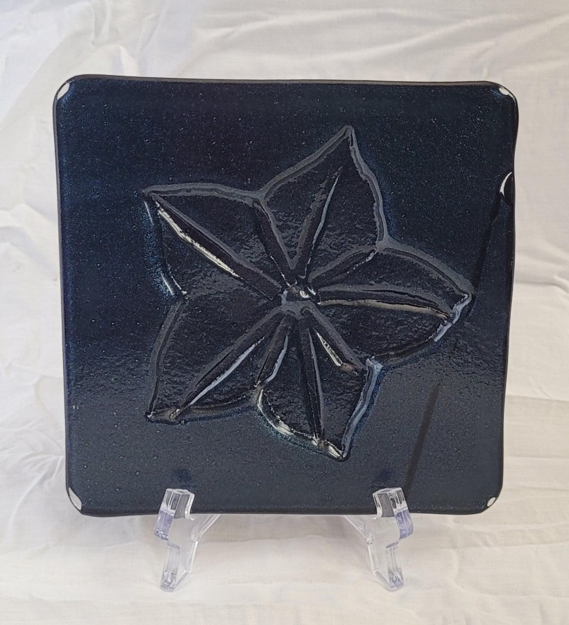 A kiln carved piece of fused glass. A design is cut out on thick fiber paper - in this case, a five pointed balloon flower. The glass is then placed over the fiber paper and fired. This produces an indentation on the glass in the form of the design. This piece has dark blue adventurine glass.