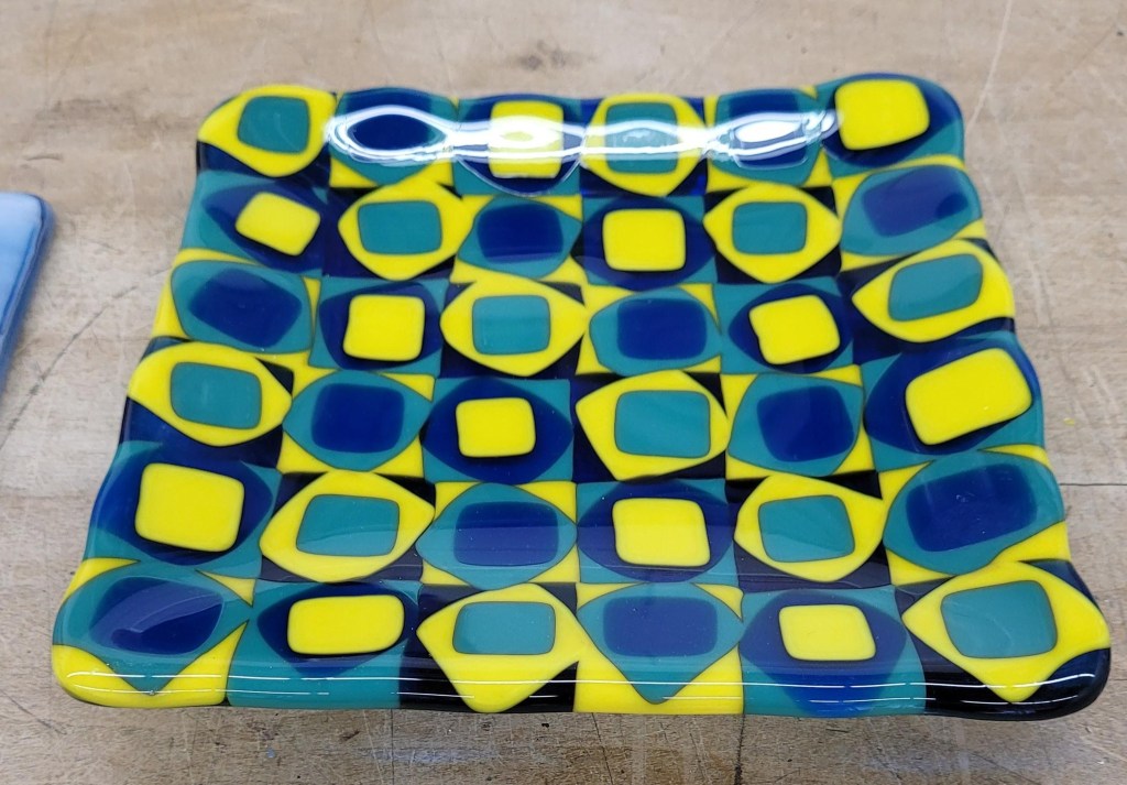 A 6x6 piece of fired fused glasswork. It was fused flat to start, then put on a mold with a square bottom and fired again so now is is raised up on the sides. It has three layers. The bottom layers are 1 inch squares, in blue, green, and yellow. The second layer is 3/4 inch squares laid on top of the 1 inch squares in a diamond shape, with yellow on blue, blue on green, and green on yellow. The third layer is 1/2 inch squares laid on the diamonds, with green on yellow, yellow on blue, and green on yellow.