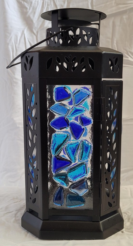 A black lantern with little cutouts, and the glass panels have fused glass. This shot has the panel with chunks of three shades of blue glass for a mosaic effect.