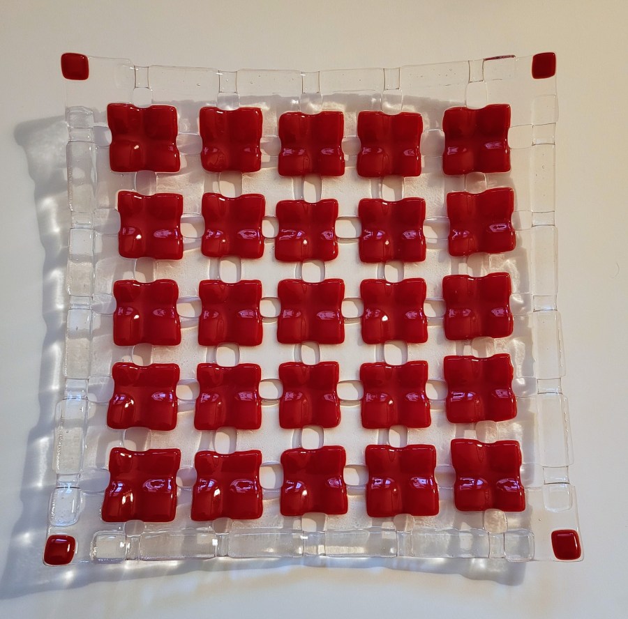 This is a fired checkerboard glass plate. It has 6 rows of 6 pieces of clear glass that are 3 by 3 centimeters, with a 1 centimeter gap in between them. On top of those squares are red glass squares, which are also 3 by 3 centimeters. They are placed so that they are covering the corners of the clear glass pieces. At each corner on the piece as a whole is a small 1 by 1 centimeter red square. It was “tack fused” which is a lower temperature fuse, and keeps a lot of the from and texture of the squares. Each red square looks a bit like a 4 petal flower - where the red corners overlapped the clear squares, they are raised up and the bits that were not overlapping the clear are sunken down a bit. After the tack fuse, there was a slump fuse where it was placed in a mold and fired again at a low temperature, and now it is gently raised on the edges.